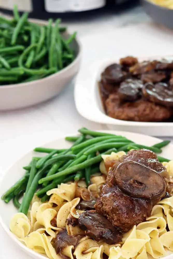 homemade slow cooker salisbury steaks on a white plate with mushrooms over egg noodles