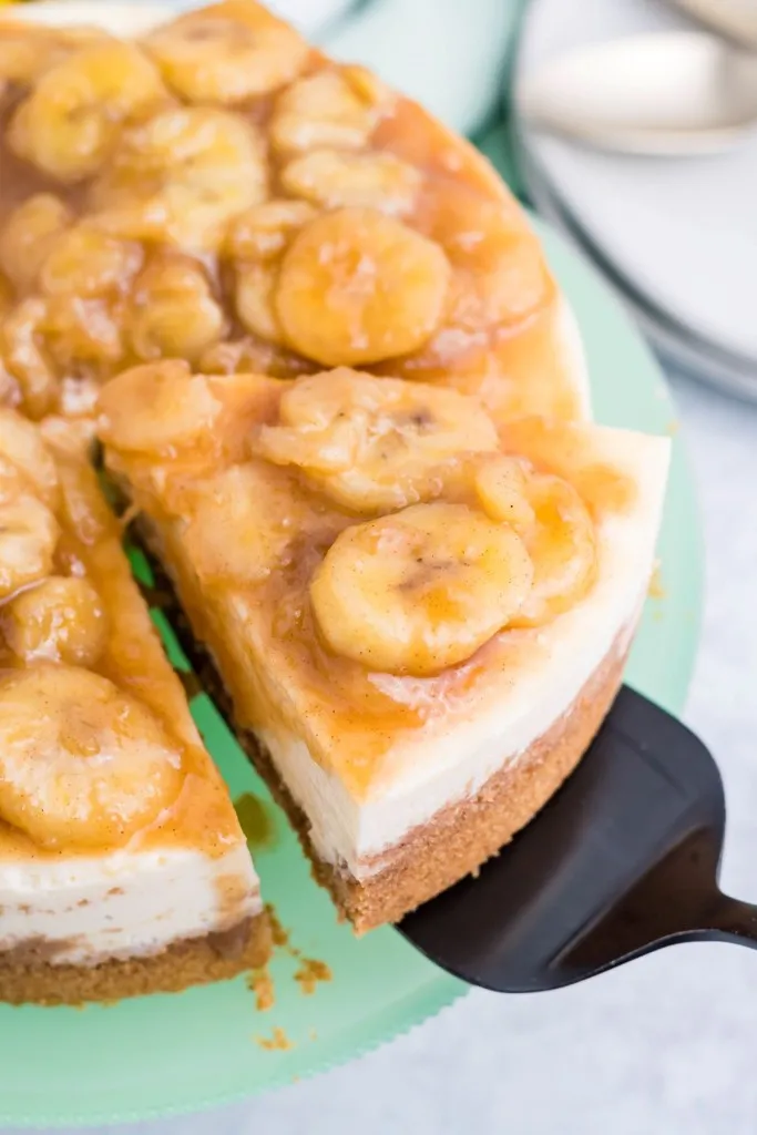 a slice of bananas foster cheesecake being life from whole cheesecake on a green platter