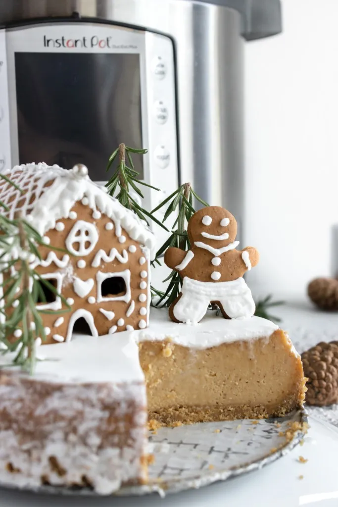 A holiday gingerbread cheesecake in front of an instant pot pressure cooker with a gingerbread cookie and house