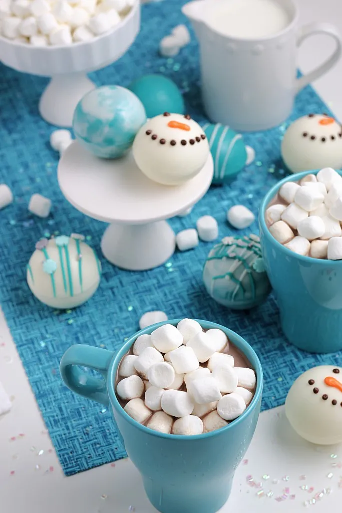 blue and white snowman hot cocoa bombs on a white pedestal and blue fabric table liner
