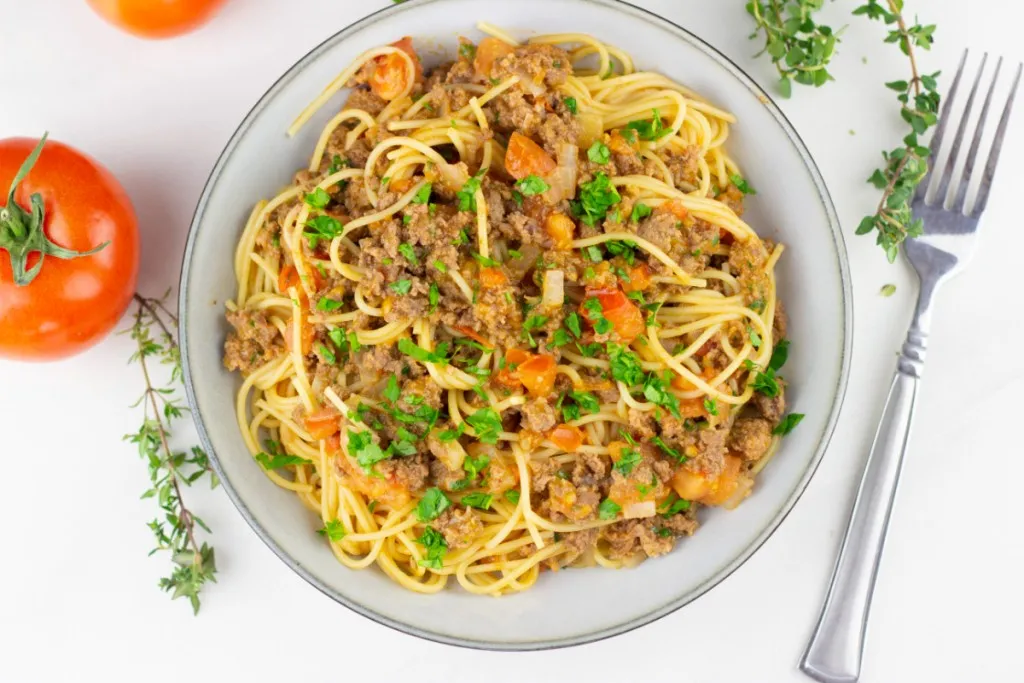 overhead flat lay view of a one pot taco spaghetti recipe with a fork and fresh red tomato in the background next ot the plate