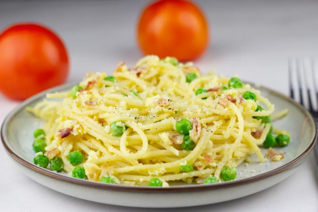 side view of pasta carbonara on a plate with a brown lip and 2 tomatoes in background
