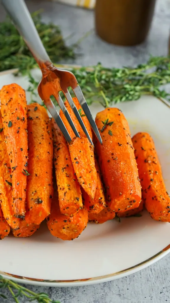 a fork stuck in a carrot. pile of herb roasted air fryer carrots and a gold trimmed white platter with fresh herbs