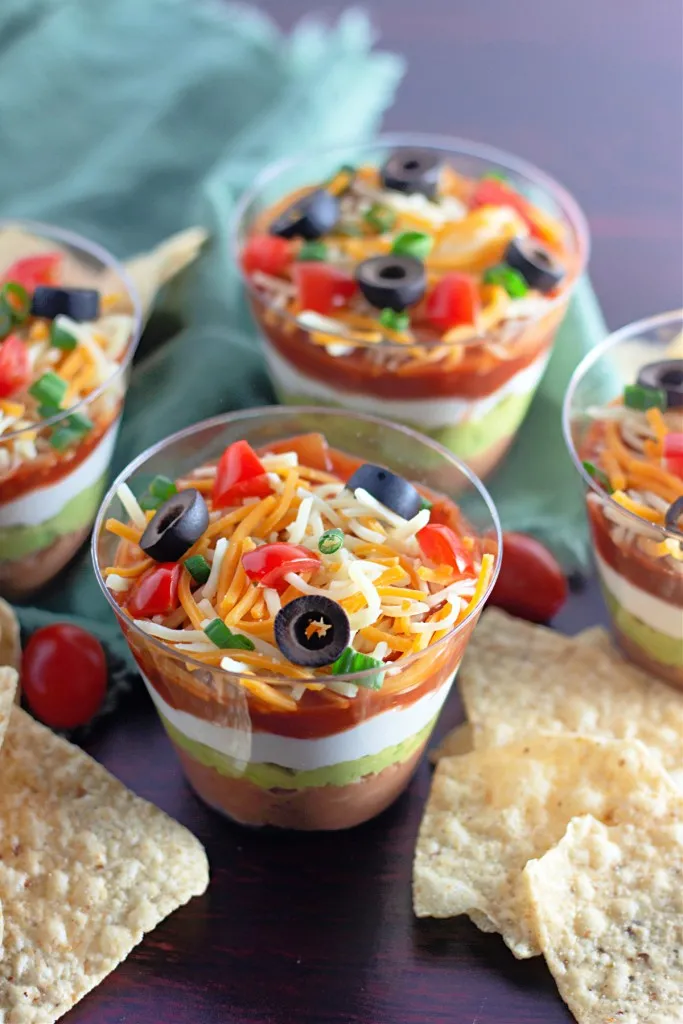 Assembled 7 Layer Dip Cups with tortilla chips and a mint kitchen cloth on a dark kitchen countertop.