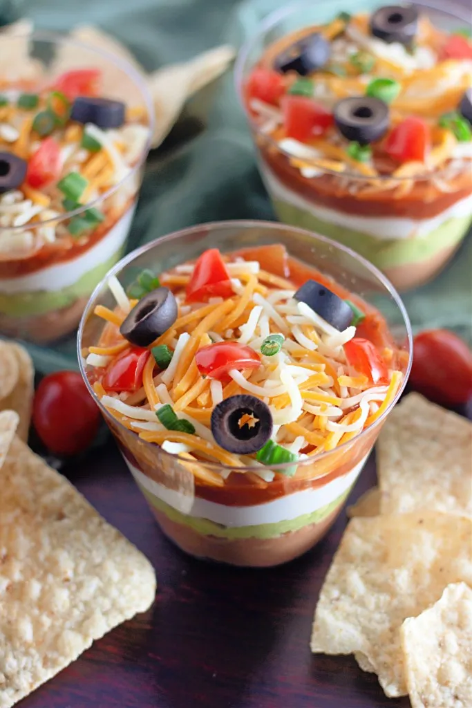 Assembled 7 Layer Dip Cups with tortilla chips and a mint kitchen cloth on a dark kitchen countertop.