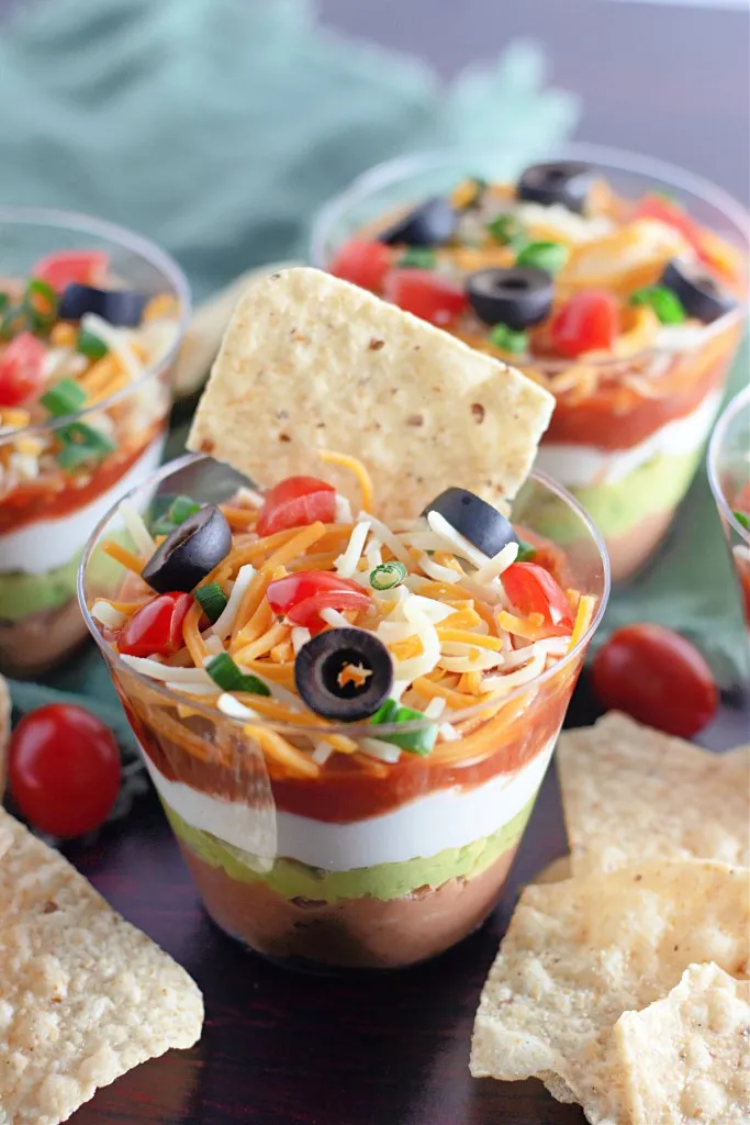Assembled 7 Layer Dip Cups with tortilla chips and a mint kitchen cloth on a dark kitchen countertop.