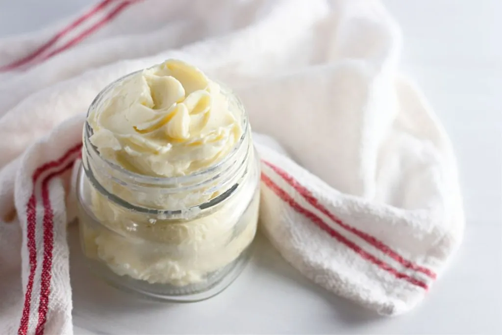 Glass jar of freshly made butter with a white a red striped kitchen cloth on a white marble countertop.