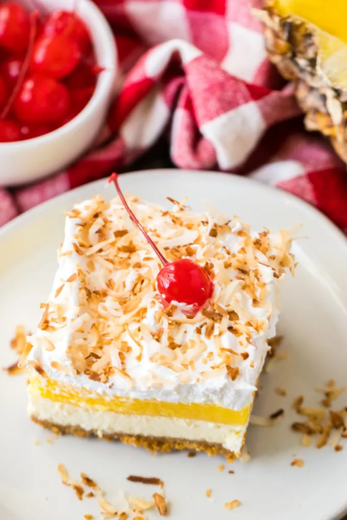 Overhead view of a slice of Pina Colada Cheesecake Bars dessert on a white serving plate, bowl of maraschino cherries, freshly cut pineapple, red and white checkered kitchen cloth.