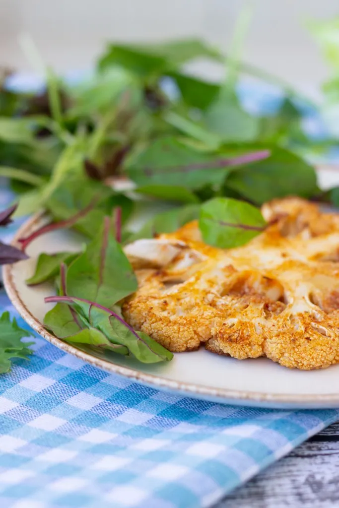 Air fryer cauliflower steak on a white serving plate with fresh greens, and white and blue table cloth.