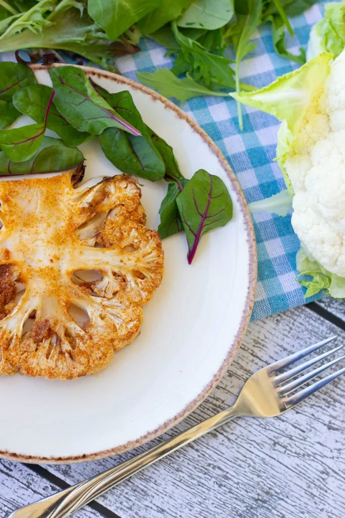 Air fryer cauliflower steak on a white serving plate with fresh greens, metal fork, head of cauliflower, and white and blue table cloth.