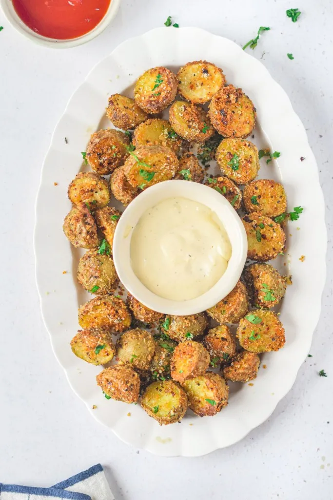 Overhead view of air fryer parmesan potatoes on a white serving plate with a small bowl of sauce in the middle, small bowl of ketchup, white kitchen cloth, on a white countertop.