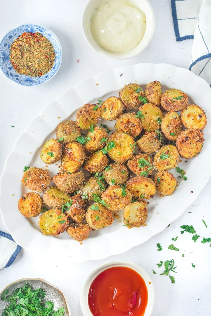 White serving plate with air fryer parmesan potatoes, bowl of ketchup, bowl of seasoning, bowl of fresh herbs, white kitchen cloth, on a white countertop.