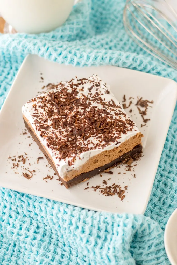 Overhead view of a slice of French Silk Brownies on a white plate, metal whisk, glass of cold milk, on a pale blue kitchen cloth.