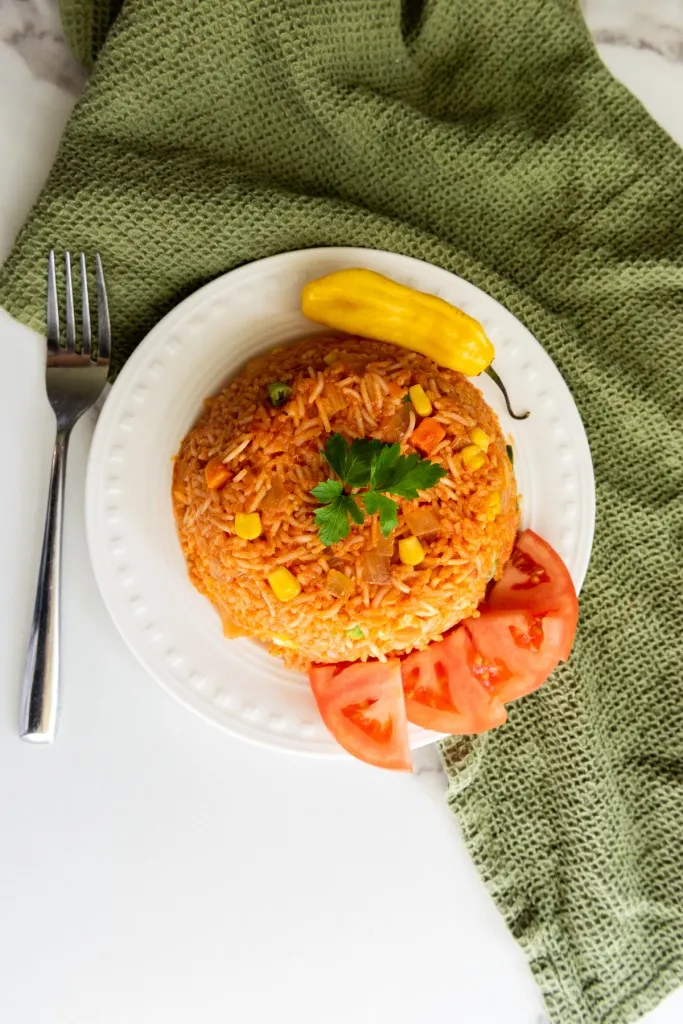 Overhead view of Jollof rice on a white serving plate with fresh tomato wedges and habanero pepper, metal fork and olive green kitchen cloth on a white marble countertop.