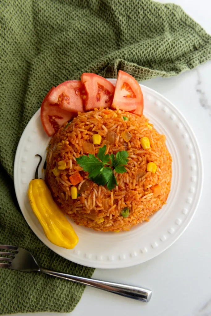 Overhead view of Jollof rice on a white serving plate with fresh tomato wedges and habanero pepper, metal fork and olive green kitchen cloth on a white marble countertop.