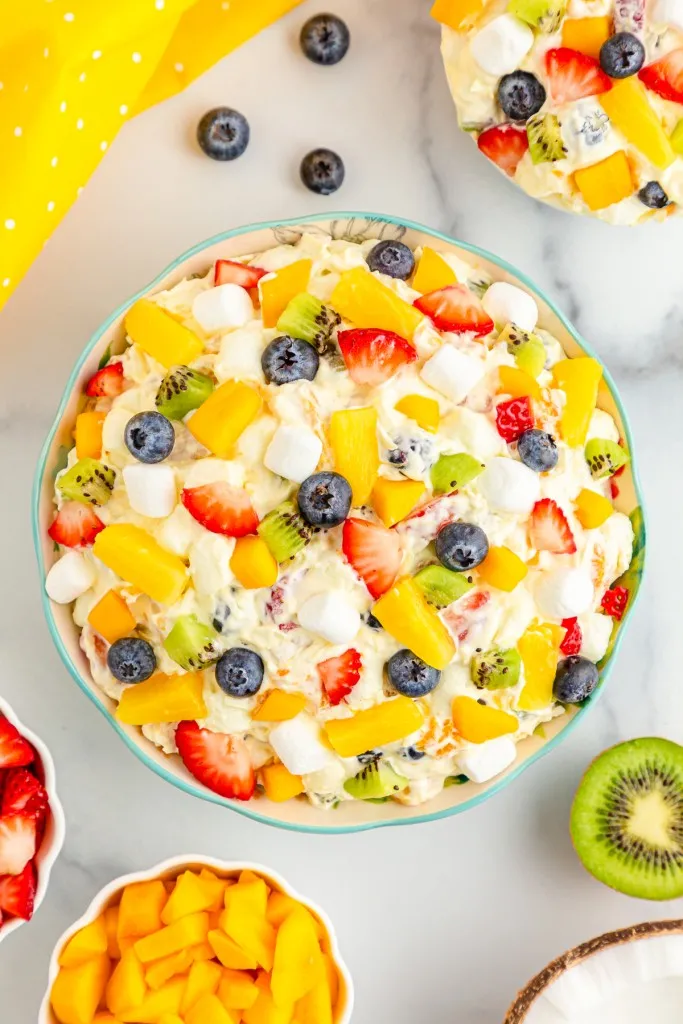 Overhead view of tropical fruit marshmallow fluff, fresh fruit, yellow kitchen cloth on a white countertop.