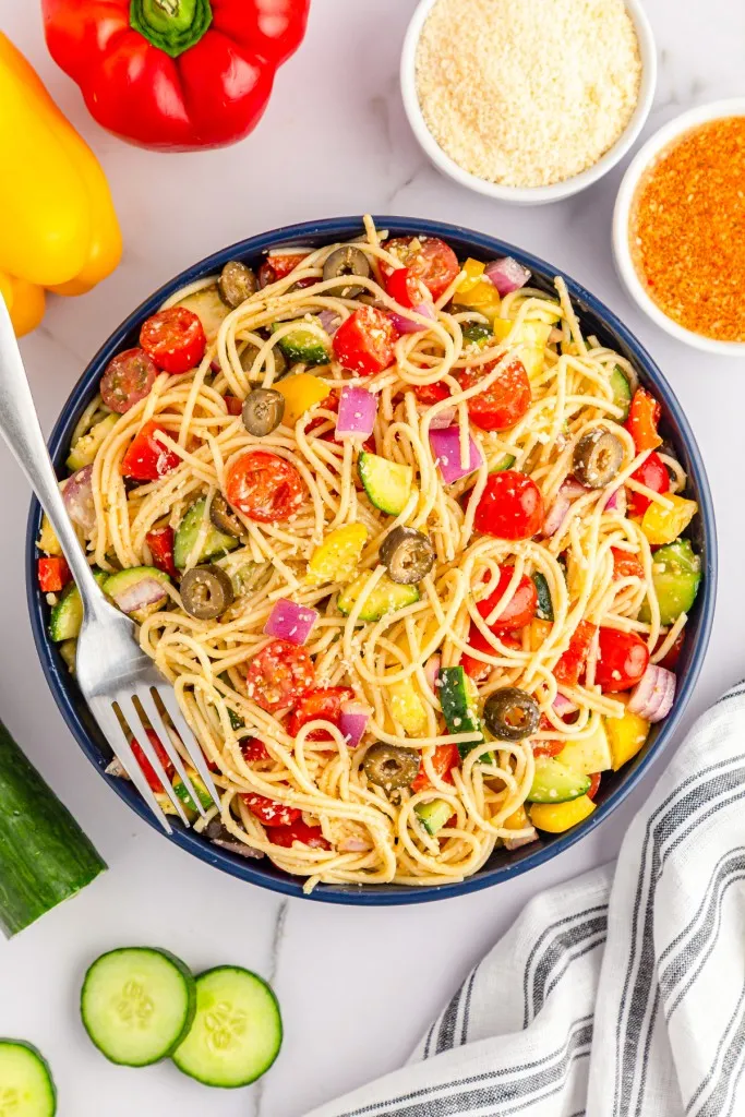 Bowl of freshly assembled California Spaghetti Salad with a metal fork, pasta salad ingredients on a kitchen countertop with a striped cloth.