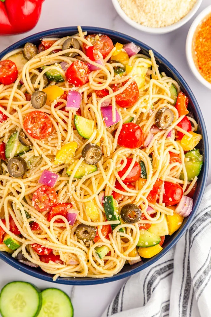 Bowl of freshly assembled California Spaghetti Salad with a metal fork, pasta salad ingredients on a kitchen countertop with a striped cloth.