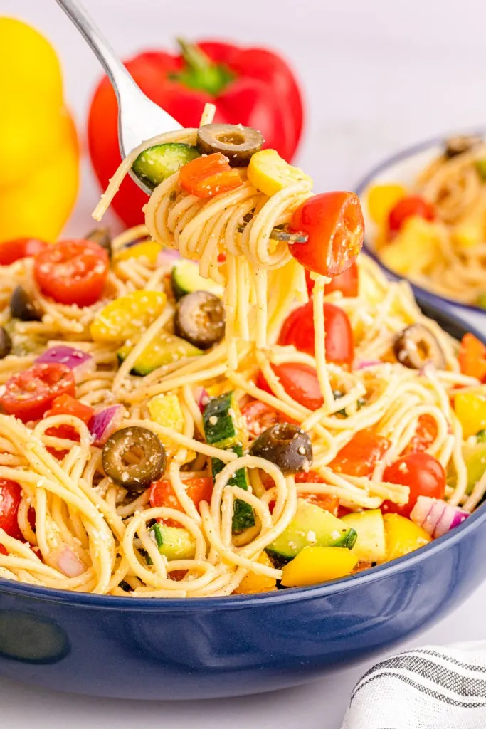 Bowl of freshly assembled California Spaghetti Salad with a metal fork twisting pasta, red and yellow bell peppers in the background with a striped kitchen cloth.