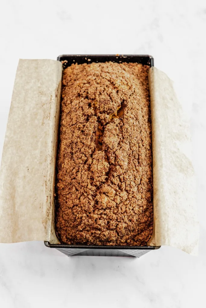 Overhead view of pumpkin streusel bread in a metal loaf pan with parchment paper hanging off both sides, on a white countertop.