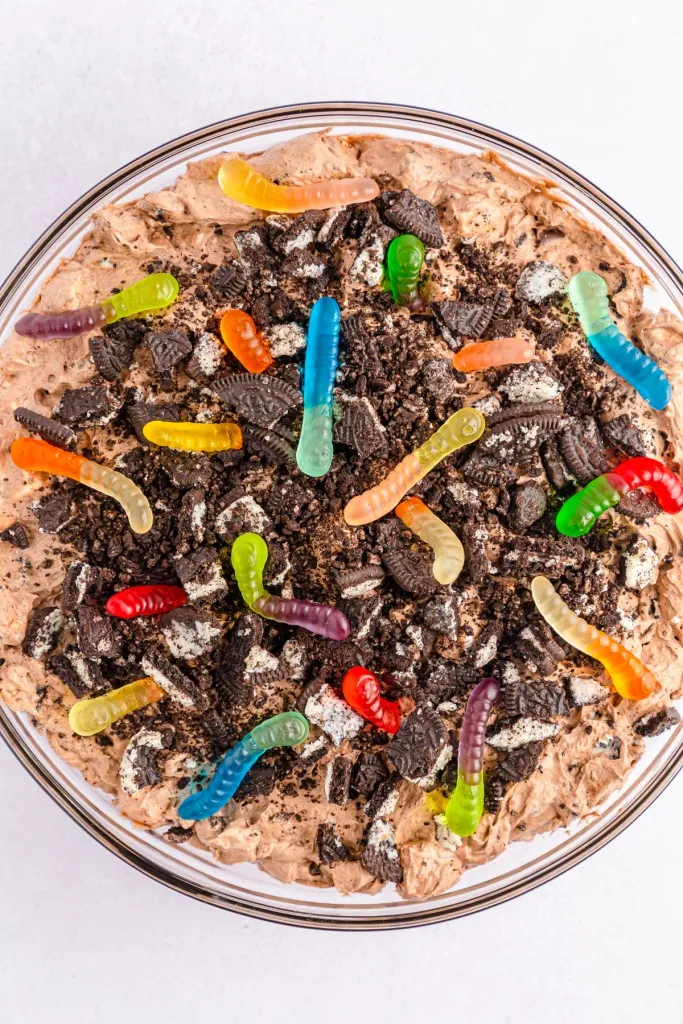 Overhead bowl of dirt worm pudding mixture topped with crushed Oreos and gummy worms on a white countertop.