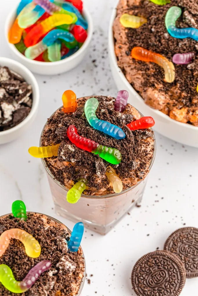Overhead view of dirt worm cups, bowl of dirt worms pudding, loose Oreos, small bowl of gummy worms and small bowl of broken Oreos, on a white countertop.