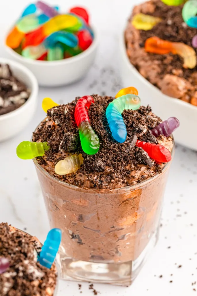 Close up of dirt worms cup, bowl of dirt worm pudding mixture, small bowls of gummy worms and broken Oreos, on a white countertop.