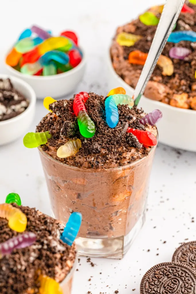 Dirt worm cups in single glasses with crushed Oreos and gummy worms, bowl of chocolate pudding with crushed Oreos and gummy worms, on a white countertop.