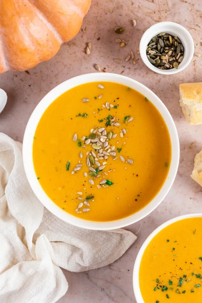 Overhead view of two bowls of pumpkin soup, whole pumpkin, small bowl of spice, crusty bread rolls, cream kitchen towel on a countertop.