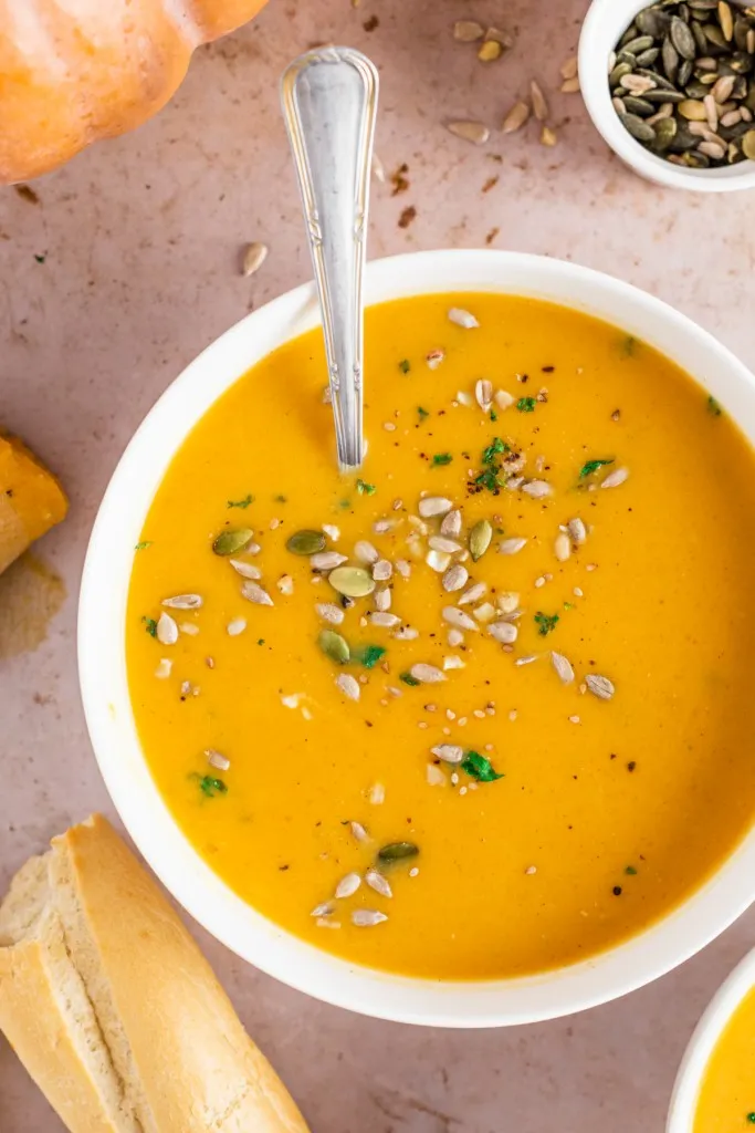 Bowl of easy pumpkin soup recipe with a metal spoon, bread roll and recipe ingredients on the countertop.