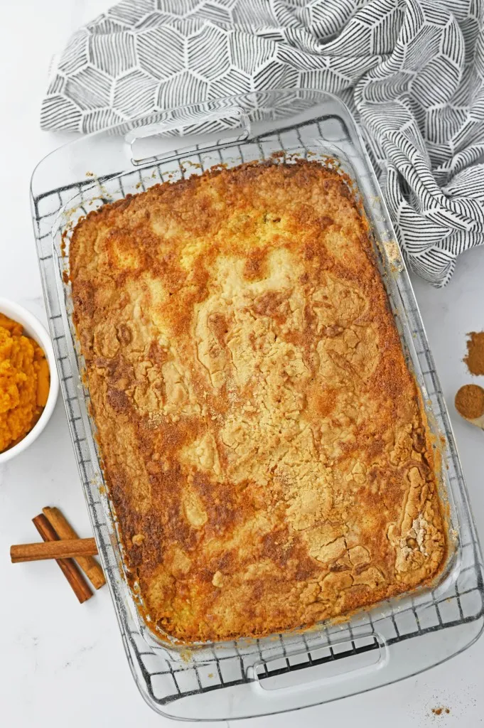 Overhead view of freshly baked pumpkin dump cake in a glass baking dish on a wire cooling rack with decorative cloth and recipe ingredients.