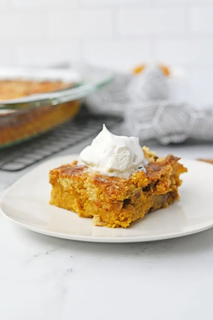 Slice of pumpkin dump cake on a white serving plate, remaining pumpkin dump cake in a glass baking dish in the background.