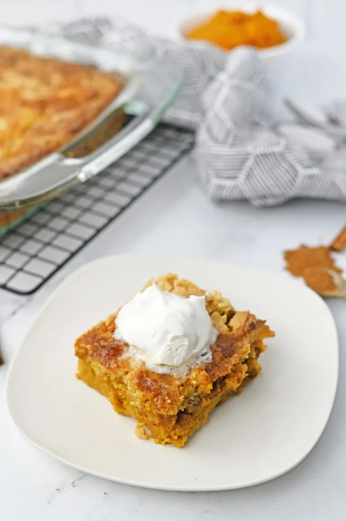 Slice of pumpkin dump cake with a dollop of Cool Whip on a white serving plate, baking dish with pumpkin dump cake on a wire cooling wrack, on a white countertop.