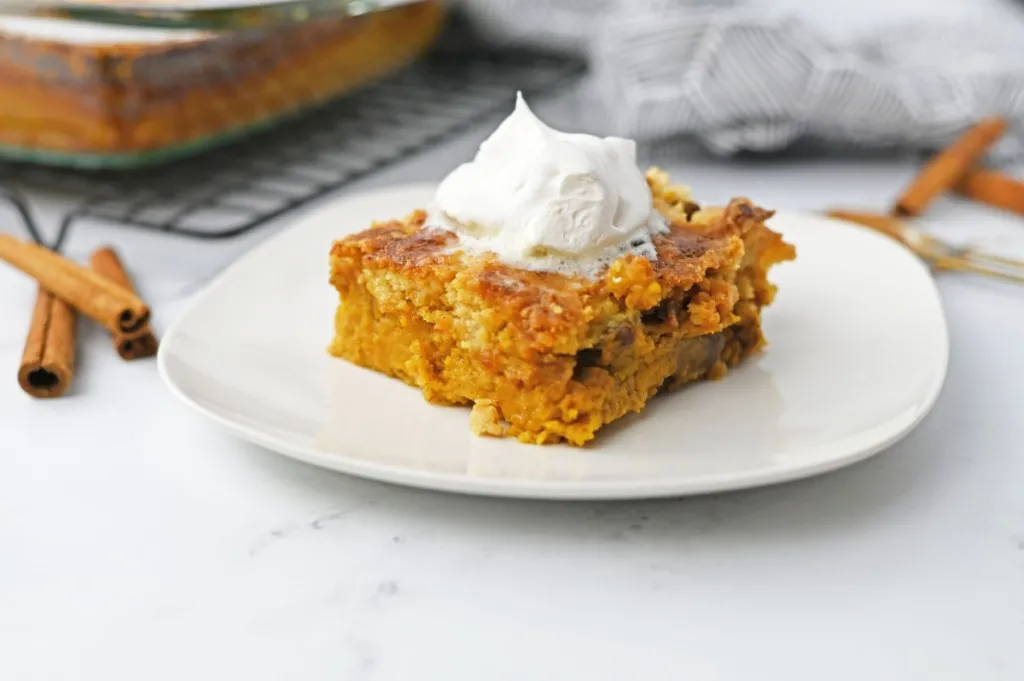 Slice of pumpkin dump cake with a dollop of Cool Whip on a white serving plate on a white countertop with remaining dump cake in the background.