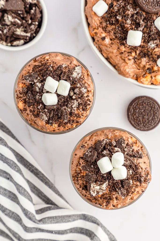 Overhead view of Oreo Fluff Salad in single glasses and larger bowl, striped kitchen towel, whole Oreos on a marble countertop.