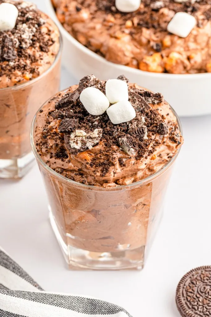 Close up of Oreo Fluff Salad in glasses, large bowl of Oreo Fluff Salad in the background, striped kitchen towel, whole Oreos on a white marble countertop.