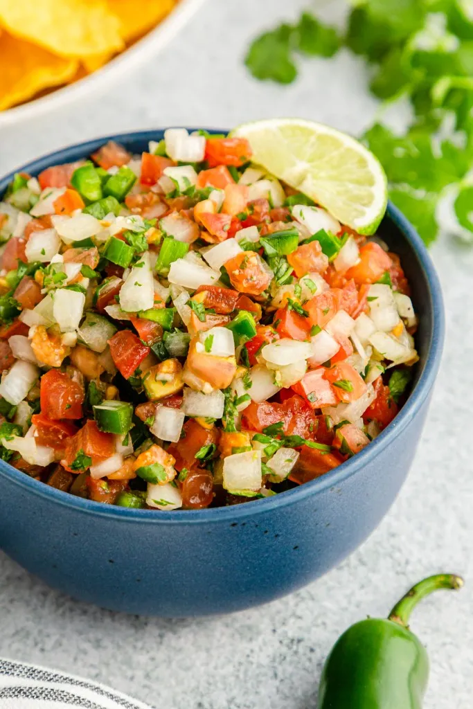 Bowl of Pico de Gallo, bowl of tortilla chips, striped kitchen cloth, jalapeno pepper.