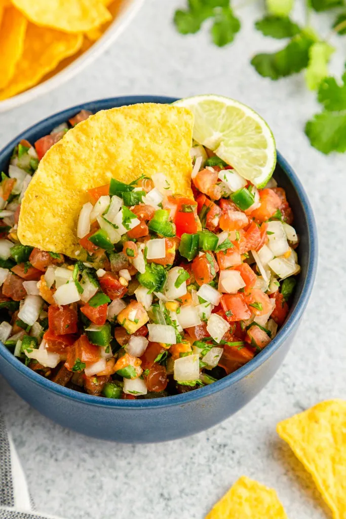 Bowl of Pico de Gallow with tortilla chip and sliced lime, bowl of tortilla chips, striped kitchen cloth, on a marble countertop.
