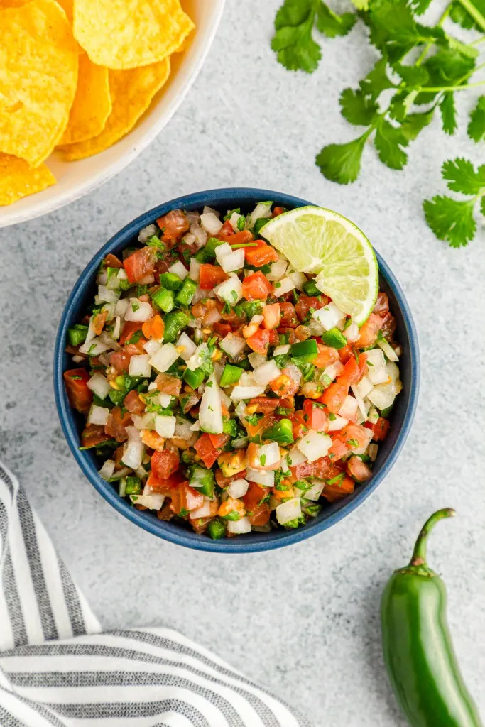 Overhead view of a bowl of Pico de Gallo, bowl of tortilla chips, striped kitchen towel, jalapeno pepper, on a marble countertop.