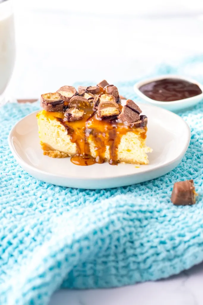 A slice of Twix cheesecake on a white serving plate on a blue kitchen cloth, chocolate syrup in a small container in the background.