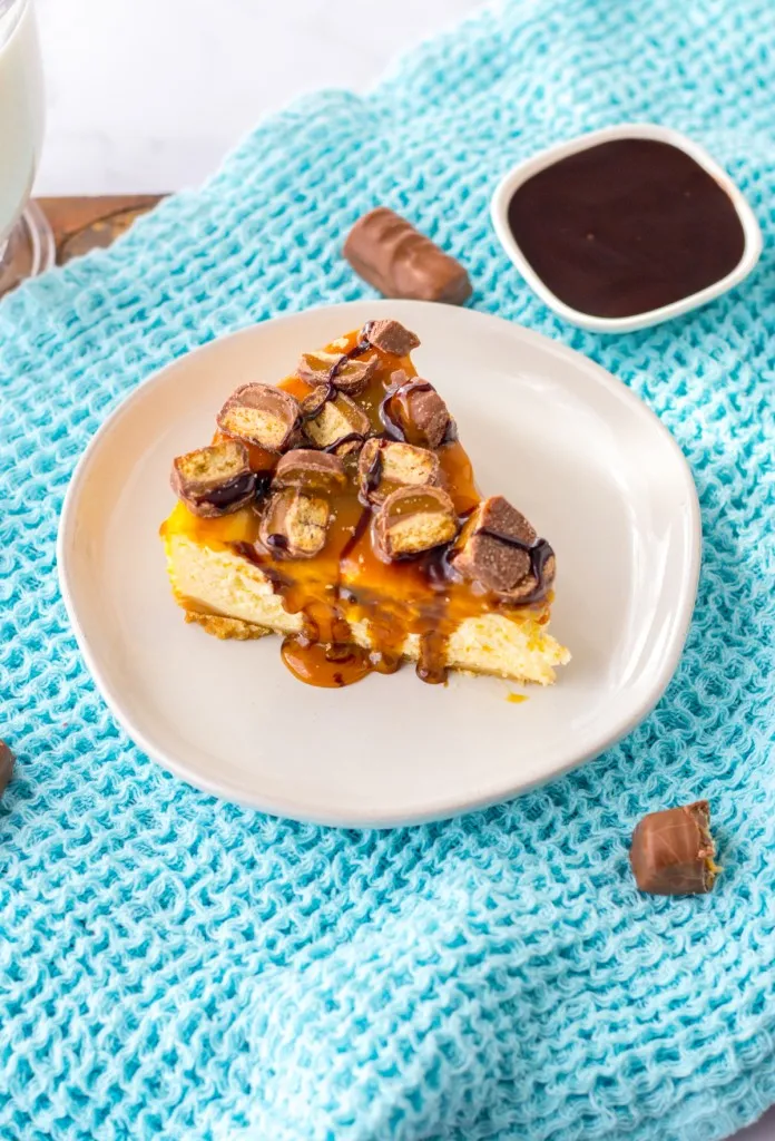 Slice of Twix cheesecake on a white plate with a light blue table cloth, bowl of chocolate syrup and broken Twix candy in the background.