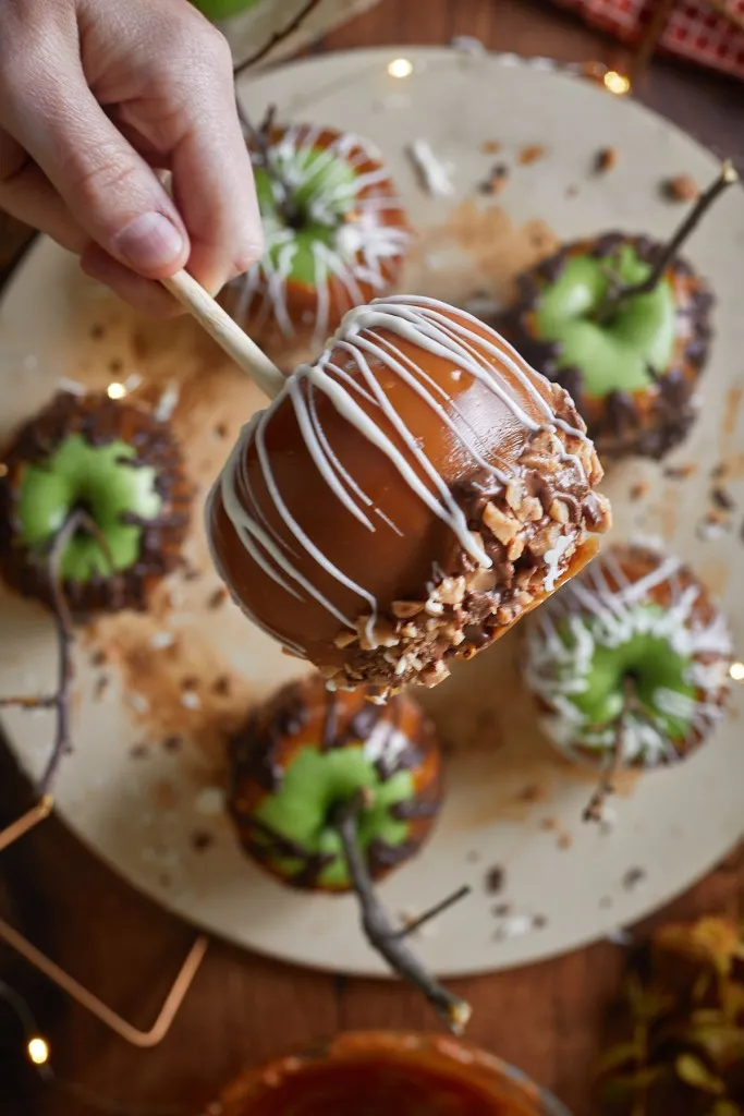 hand holding a caramel apple above a large serving board with more caramel apples.