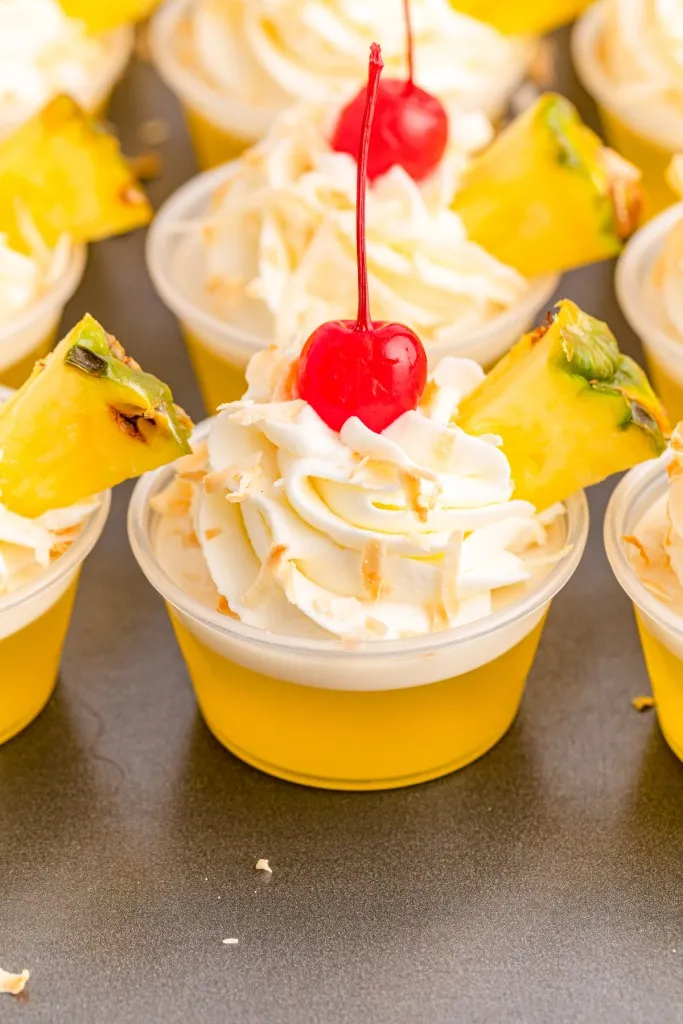 Assembled Pina Colada Jello Shots on a countertop with whipped cream, maraschino cherries, and pineapple wedges.