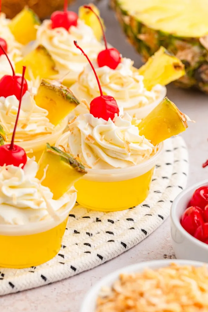 Pina colada jello shots on a decorative mat surrounded by recipe ingredients on a white countertop.