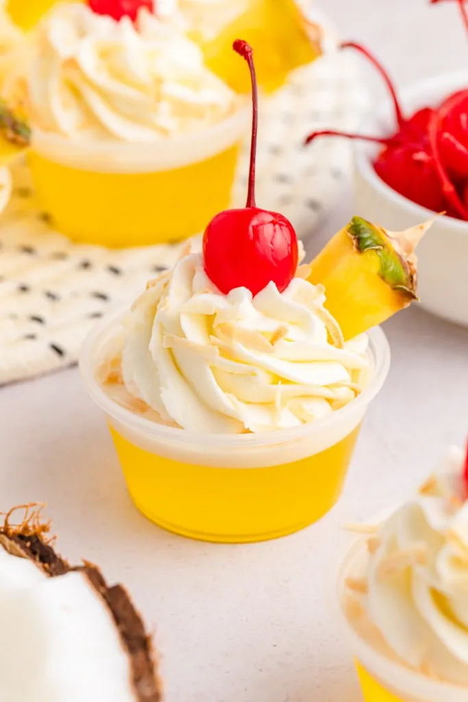 Close up of Pina Colada Jello shots, bowl of maraschino cherries, cracked coconut on a white countertop.
