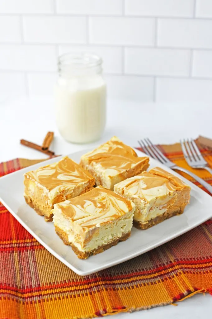 Pumpkin cheesecake bars on a white plate, glass of milk, orange checkered table cloth.
