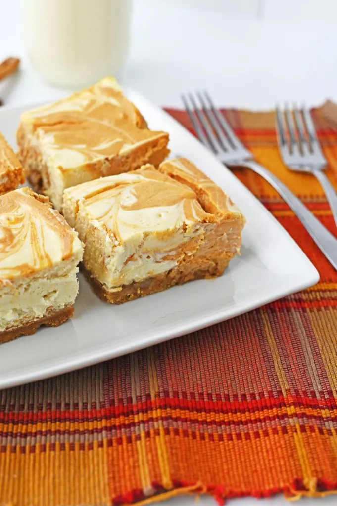 Close up of pumpkin cheesecake bars on a white plate, glass of milk, orange checkered cloth.