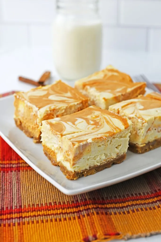 Pumpkin cheesecake bars on a white plate, glass of milk, on an orange checkered cloth.