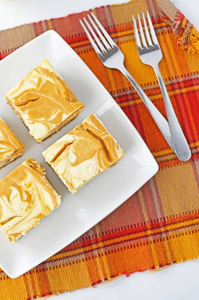 Overhead view of cheesecake bars on a white plate, two metal forks, orange checkered cloth.