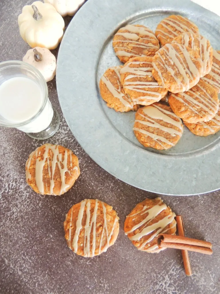 Pumpkin oatmeal cookies on a grey plate, additional cookies scattered around the plate, glass of milk, white kitchen towel on a wooden surface.